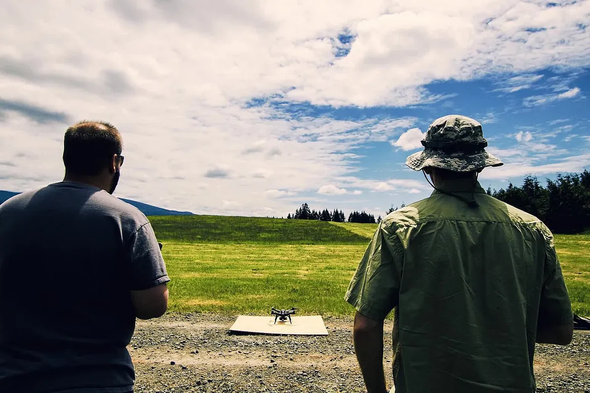 Two people face a small quadcopter on a flat launch pad in an open grass field during drone training.