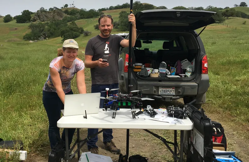 A woman and a man stand beside a folding table with a small quadcopter and GPS equipment in a grassy field.